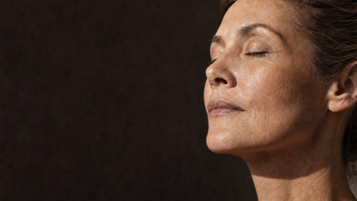 A woman with her eyes closed, face tilted towards warm light against a dark background