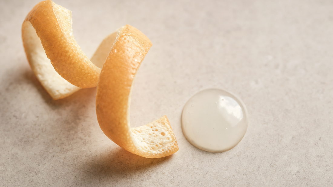 Two curls of orange peel beside a clear droplet of serum on a pale stone surface