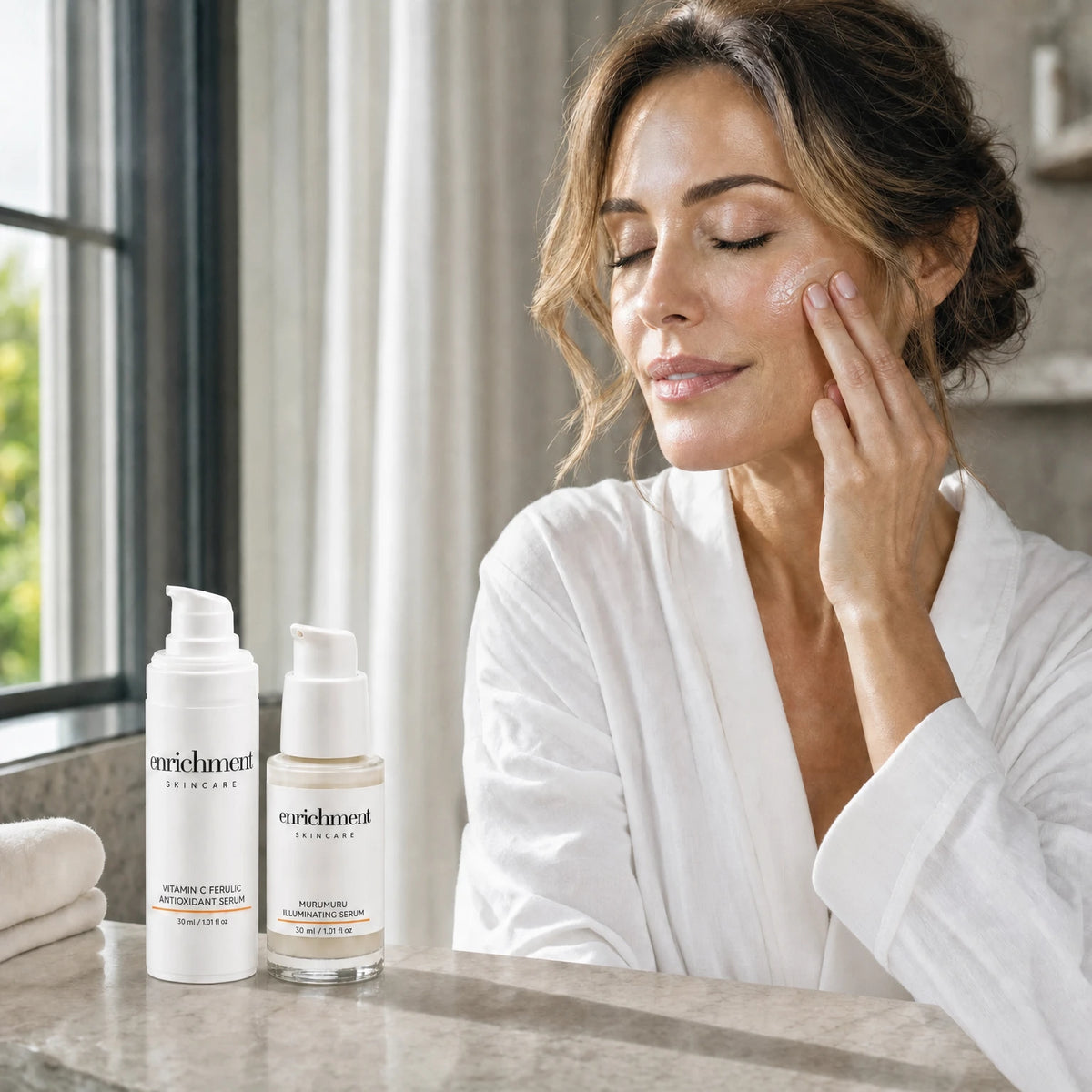 A woman in her forties applying serum to her cheek in morning light, with the two serums of the Duo on a stone washstand beside her