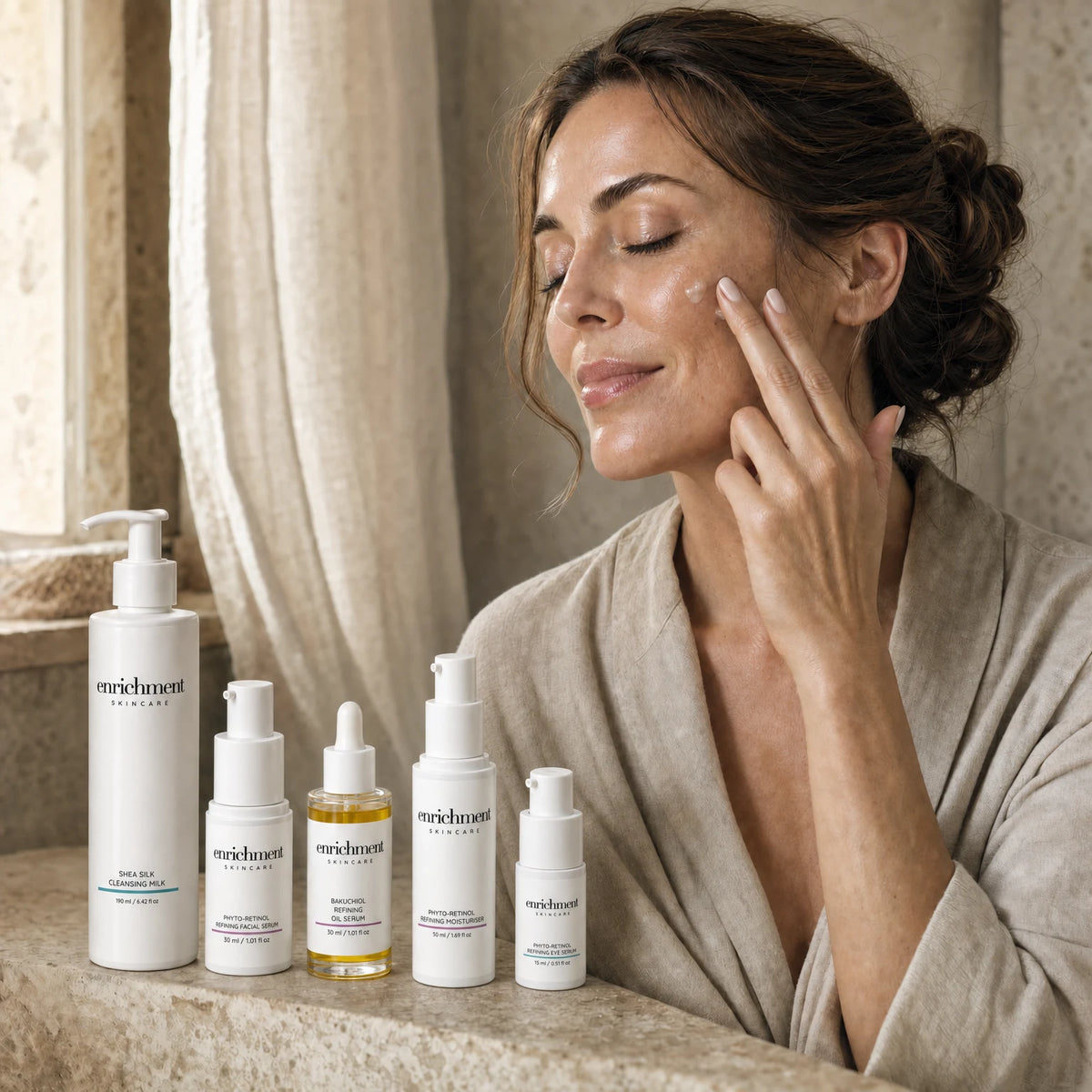 A woman in her forties applying serum to her cheek, with the five products of the set on a stone shelf in front of her