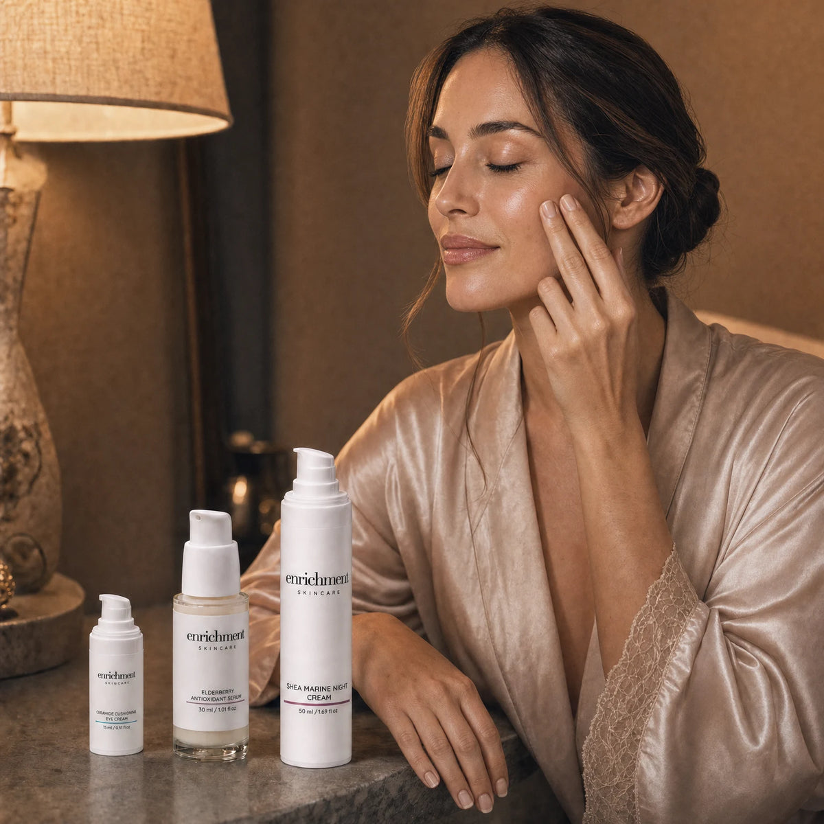 A young woman in a silk robe at a stone dressing table in warm evening lamplight, gently applying a product to her cheek with two fingertips, with Ceramide Cushioning Eye Cream, Elderberry Antioxidant Serum and Shea Marine Night Cream in the foreground
