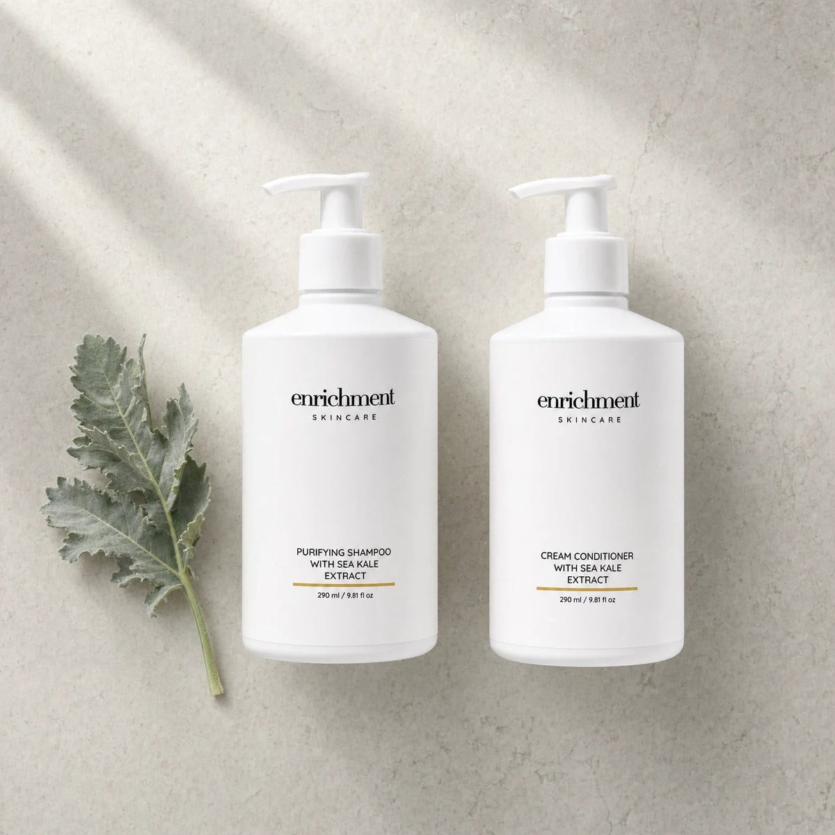 The shampoo and conditioner laid side by side on a pale stone surface with a sprig of silvery-green leaves