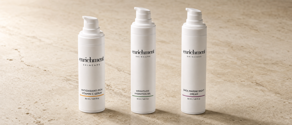 Enrichment Skincare Antioxidant-Rich Vitamin C Serum, Weightless Hydration Gel and Shea Marine Night Cream standing together on a warm travertine stone surface in soft morning light.