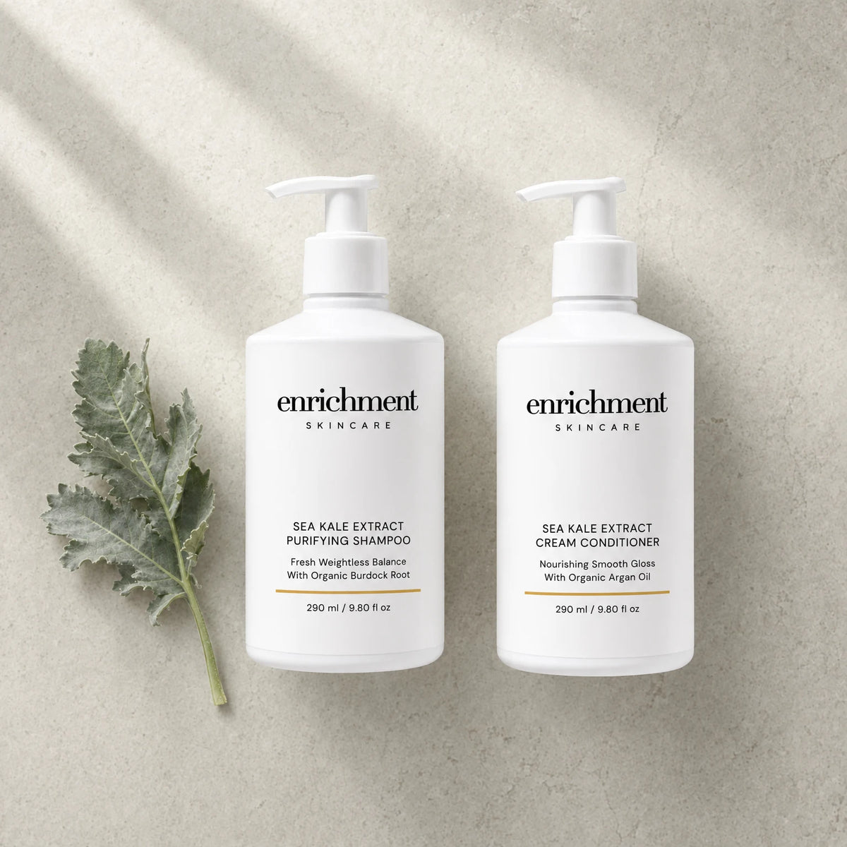 The shampoo and conditioner laid side by side on a pale stone surface with a sprig of silvery-green leaves