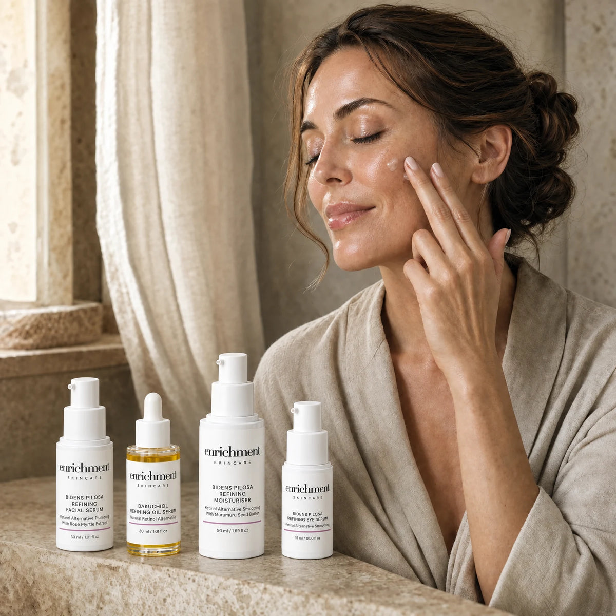A woman in her forties applying serum to her cheek, with the four products of the set on a stone shelf in front of her